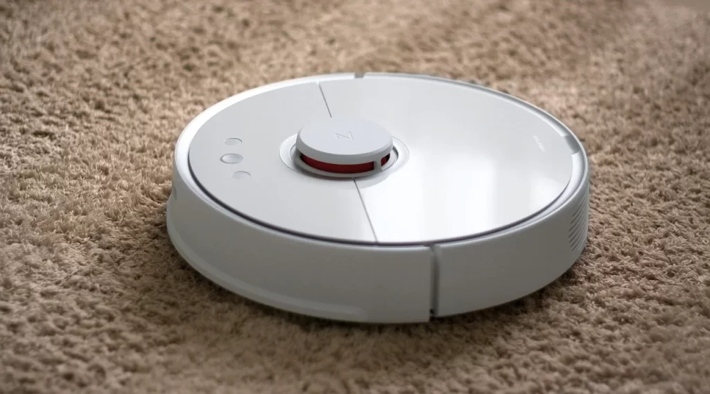 Robot pembersih lantai otomatis berwarna putih sedang membersihkan karpet berbulu di dalam ruangan rumah